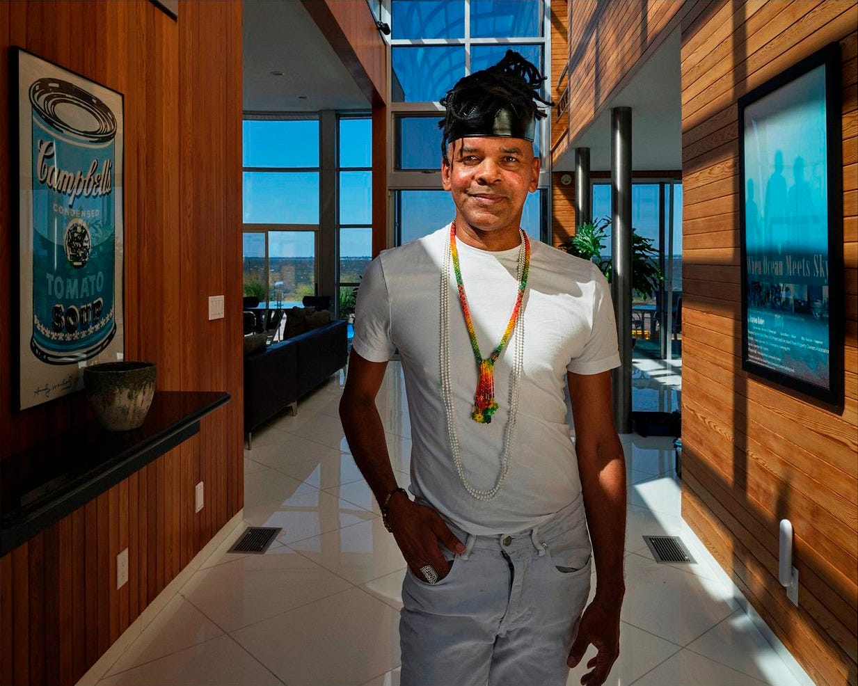 A black man in a white t-shirt and long necklaces stands in a sunlit modern hallway, with art on wooden walls and a city view in background, conveying relaxed confidence.He has his right hand resting on his light blue jean pocket, and he is staring straight into the camera. He has braids situated in a bun on top of his head, held together by a black bandana. A black man in a white t-shirt and long necklaces stands in a sunlit modern hallway, with art on wooden walls and a city view in background, conveying relaxed confidence.He has his right hand resting on his light blue jean pocket, and he is staring straight into the camera. He has braids situated in a bun on top of his head, held together by a black bandana.