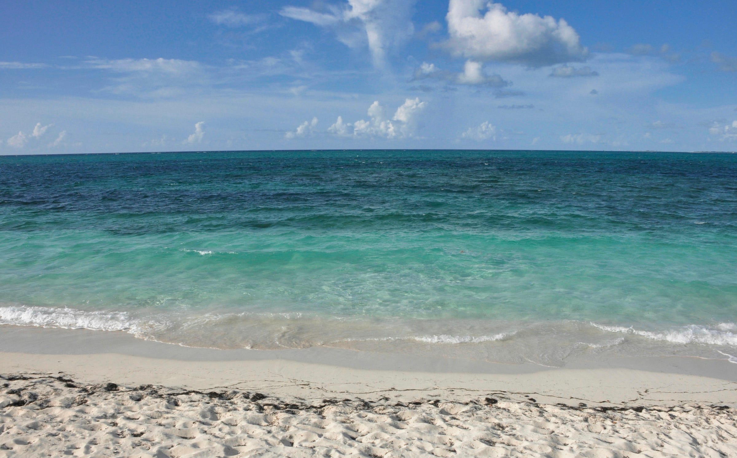 A view of waves rolling onto a sandy beach and the turquise water deepens in color as you look on the horizon where the blue sky and white cloiuds meet