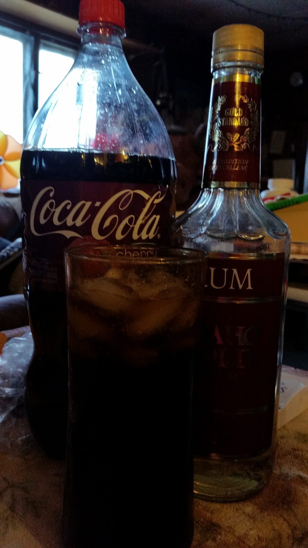 A glass of cocktail with ice cubes sits next to a bottle of cherry-flavored cola and a bottle of clear rum. The scene is dimly lit, suggesting a cozy atmosphere.