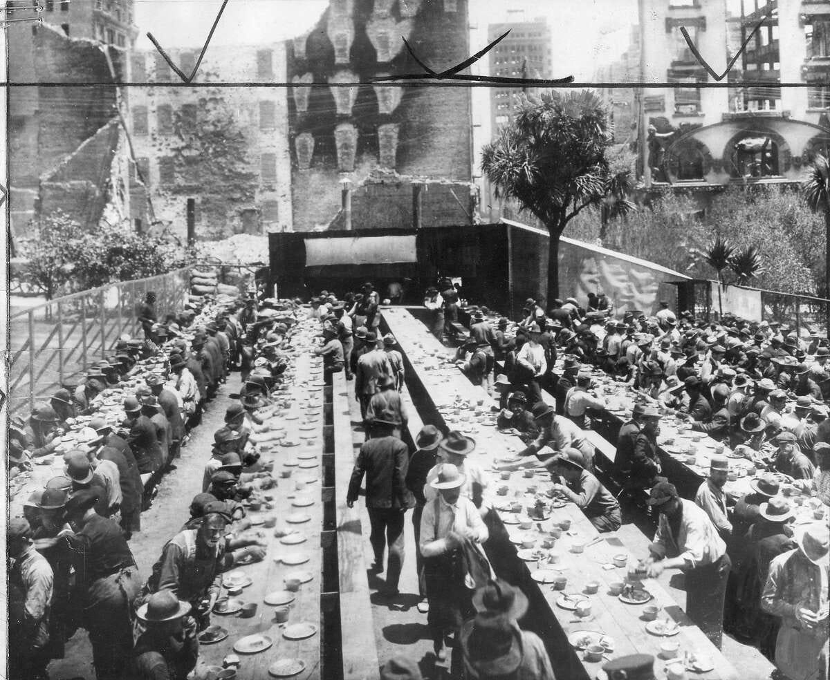 An old black and white photo shows several rows of long tables with dozens of people seated and eating meals, with the backdrop of burned out brick buildings and other devastation from the 1906 San Francisco earthquake.