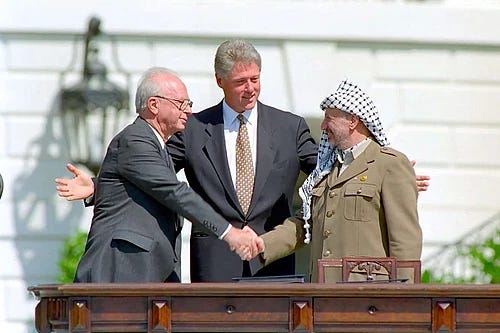 Bill Clinton with his arms behind two men who are shaking hands, as if he's about to hug them