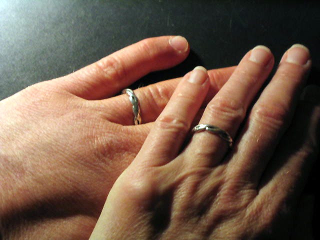 Closeup of two left hands, one on top of the other, matching silver bands on their ring fingers