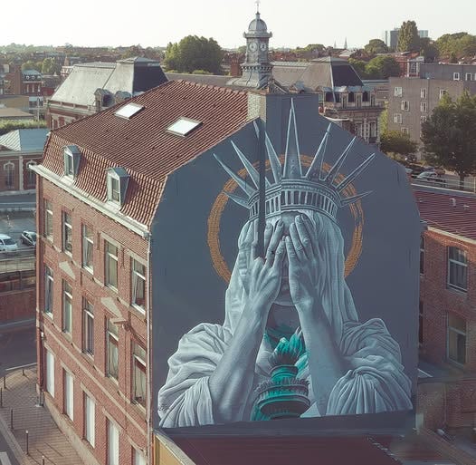 Mural by JDL Street Art in Roubaix, Lille, France, depicting the Statue of Liberty covering her face with both hands in a gesture of shame. The figure is painted in shades of grey with teal accents on the torch and crown, set against a muted background with a golden halo-like circle. The artwork spans the side of a tall brick building, contrasting modern street art with historic European architecture in the surrounding cityscape.