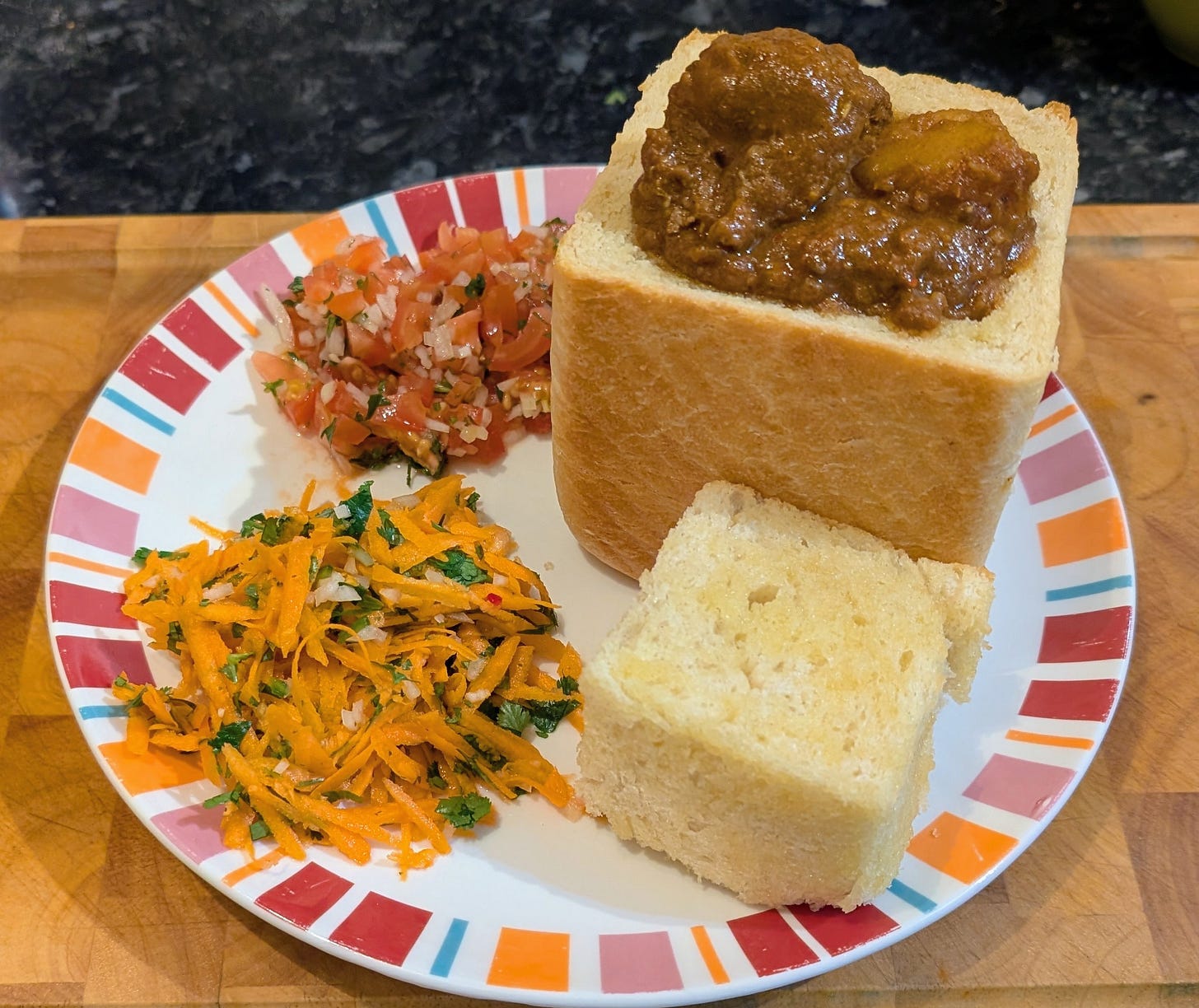 A hollowed-out loaf of white bread filled with a rich, brown South African curry sits on a patterned plate. The removed bread “lid” rests beside it. The curry contains chunks of meat and potato in a thick gravy. On the side are two fresh salads: a bright carrot-and-herb mix and a finely chopped tomato–onion sambal. The plate rests on a wooden surface.