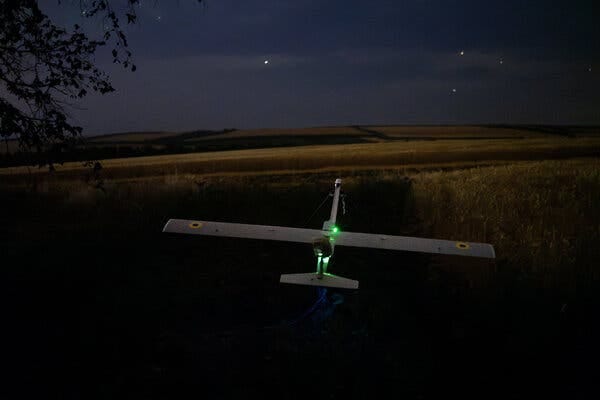 A small drone, lit by green and blue lights, sits in dark grass. Golden fields and distant hills are under a night sky with a handful of stars.