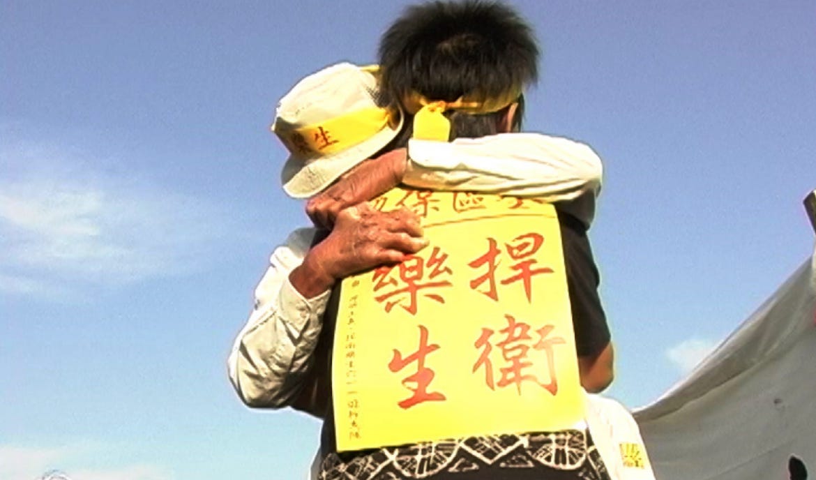 A young figure stands with their back to the camera, wearing a yellow protest sign. They are embraced by a figure with gnarled, swollen hands and wearing a khaki bucket hat