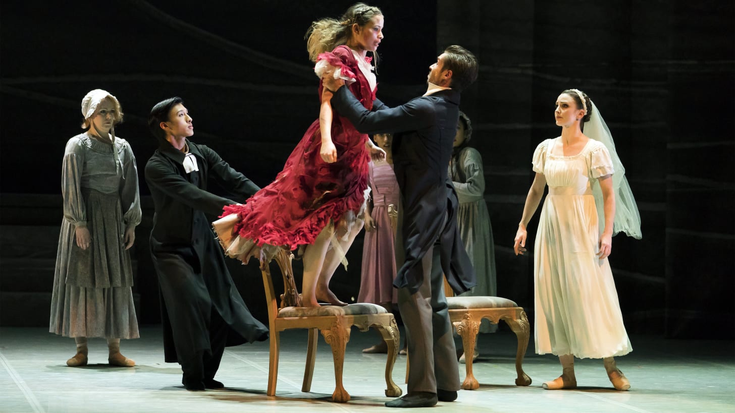 A male dancer in mid-Victorian dress holds a woman in a flame-red dress who is standing on a chair whilst a bride looks on.