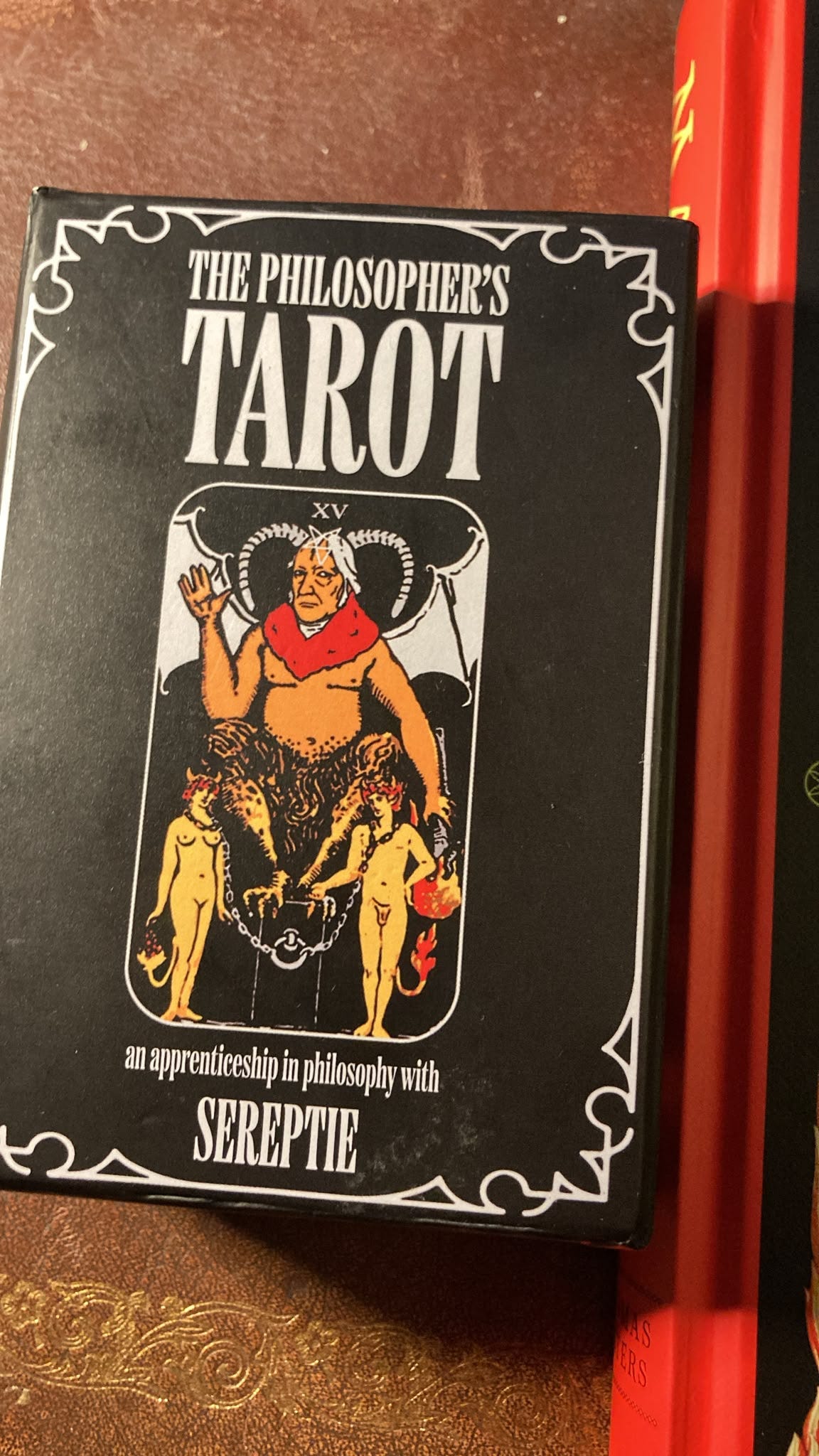 Black box of The Philosopher’s Tarot by Sereptie, featuring a philosophical reinterpretation of the traditional Devil tarot card, with a horned figure and two chained figures beneath. Black box of The Philosopher’s Tarot by Sereptie, featuring a philosophical reinterpretation of the traditional Devil tarot card, with a horned figure and two chained figures beneath.