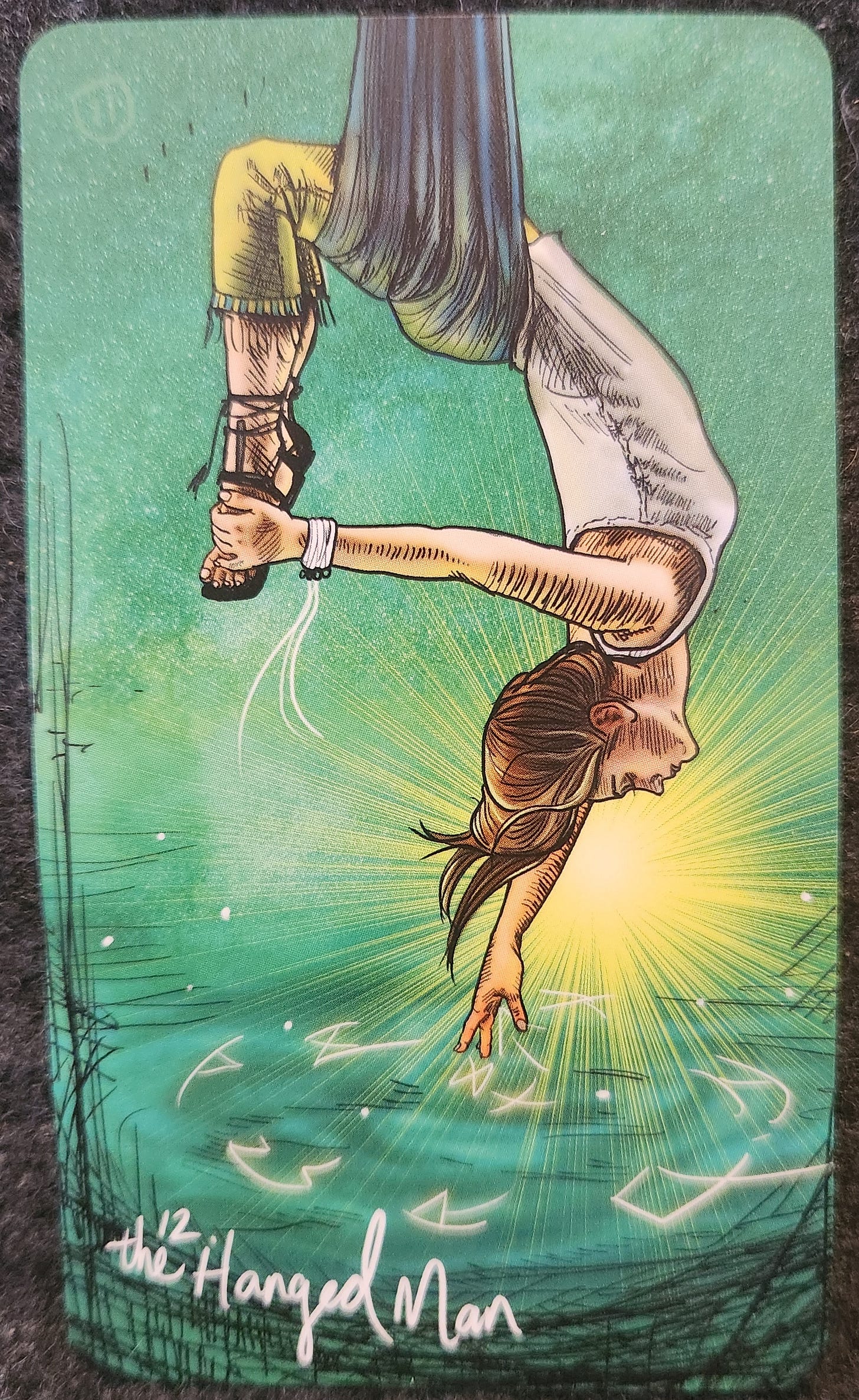 A tarot card labelled 12. The Hanged Man. A woman is hanging from a strip of aerial silk (large, thick fabric hanging from above). Her legs are draped on one side of the fabric and her torso is draped over the other. She is grasping her own ankle with one hand. Her eyes are closed and her face looks peaceful. There is a golden halo of light surrounding her head. Her other hand is pointing downwards towards the ground below, which appears to be blue-green water covered in runes. 