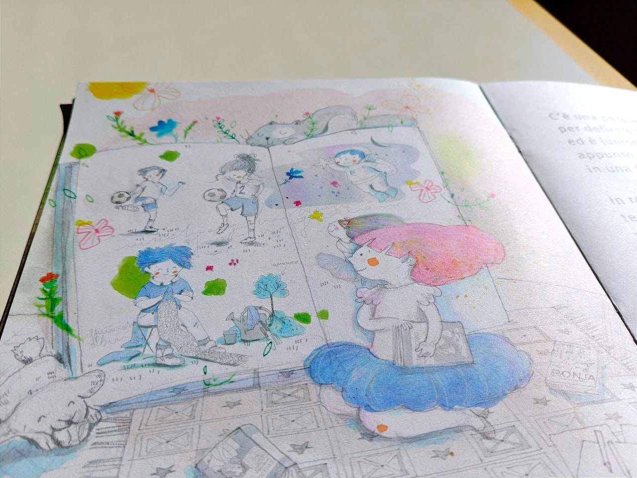 Pagina illustrata della Scatolina JUNIOR edita da effequ, appoggiata su un tavolo di legno color bianco sporco, con uno spicchio di un muro in ombra nell'angolo in alto a destra. Nel disegno compare un grande libro aperto, tenuto in verticale, con una bambine e bambini impegnati a palleggiare con un pallone da calcio, fare a maglia, esplorare lo spazio da astronauta, danzare; in primo piano, che guarda il libro, c'è una bambina coi capelli rosa che regge sottobraccio un libro. Sopra al libro gigante sonnecchia un gatto, mentre in basso un cane gioca con una pallina. La tecnica del disegno è a matita e acquerello.