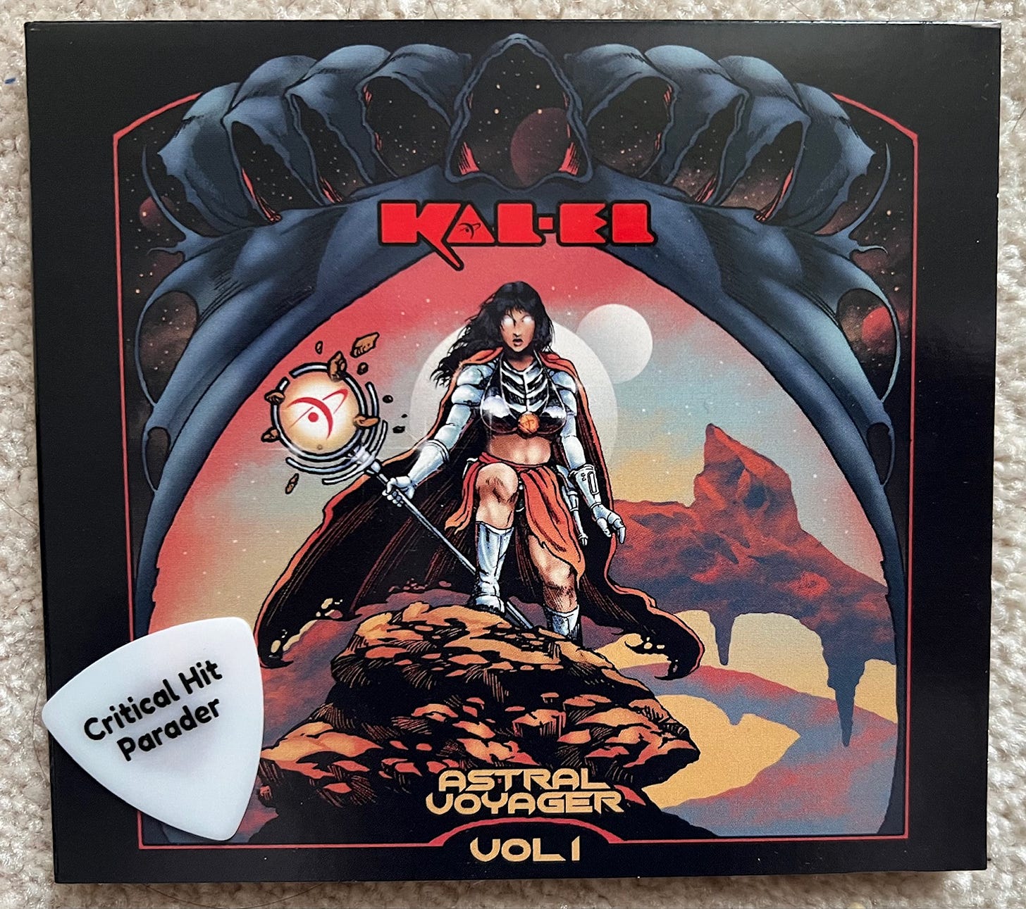 A photo of the CD "Astral Voyager Vol. 1" by Kal-Ed. If features artwork by Steven Yoyada depicting a feminine figure with glowing eyes holding a staff on a rocky planetary landscape. A white Critical Hit Parader guitar pick is laying on the bottom left corner of the CD as if in orbit.