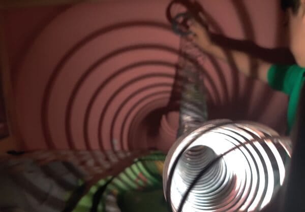 A little boy holds a slinky in front of a flashlight that is casting looping circle shadows on a pink wall in the dark.