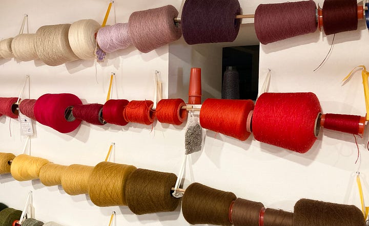 Two photographs showing firstly a wall with cones of coloured wool hung on poles and secondly a hand with two shades of red wool blended together ready for knitting.