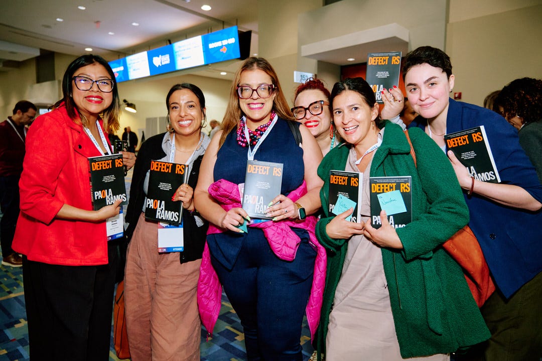 Six women in the convention center hallway all holding signed copies of Defectors by Paola Ramos pressed together smiling for the photo in a row