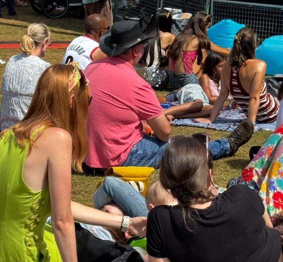 A group of people sitting on the grass at an outdoor festival. Peter is wearing a bright pink top and sunglasses, seated among others on blankets and mats. It is a sunny day with a relaxed, communal atmosphere.