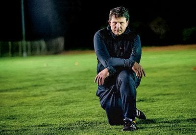 Coach Pete Sickle kneels on the grass of a rugby field