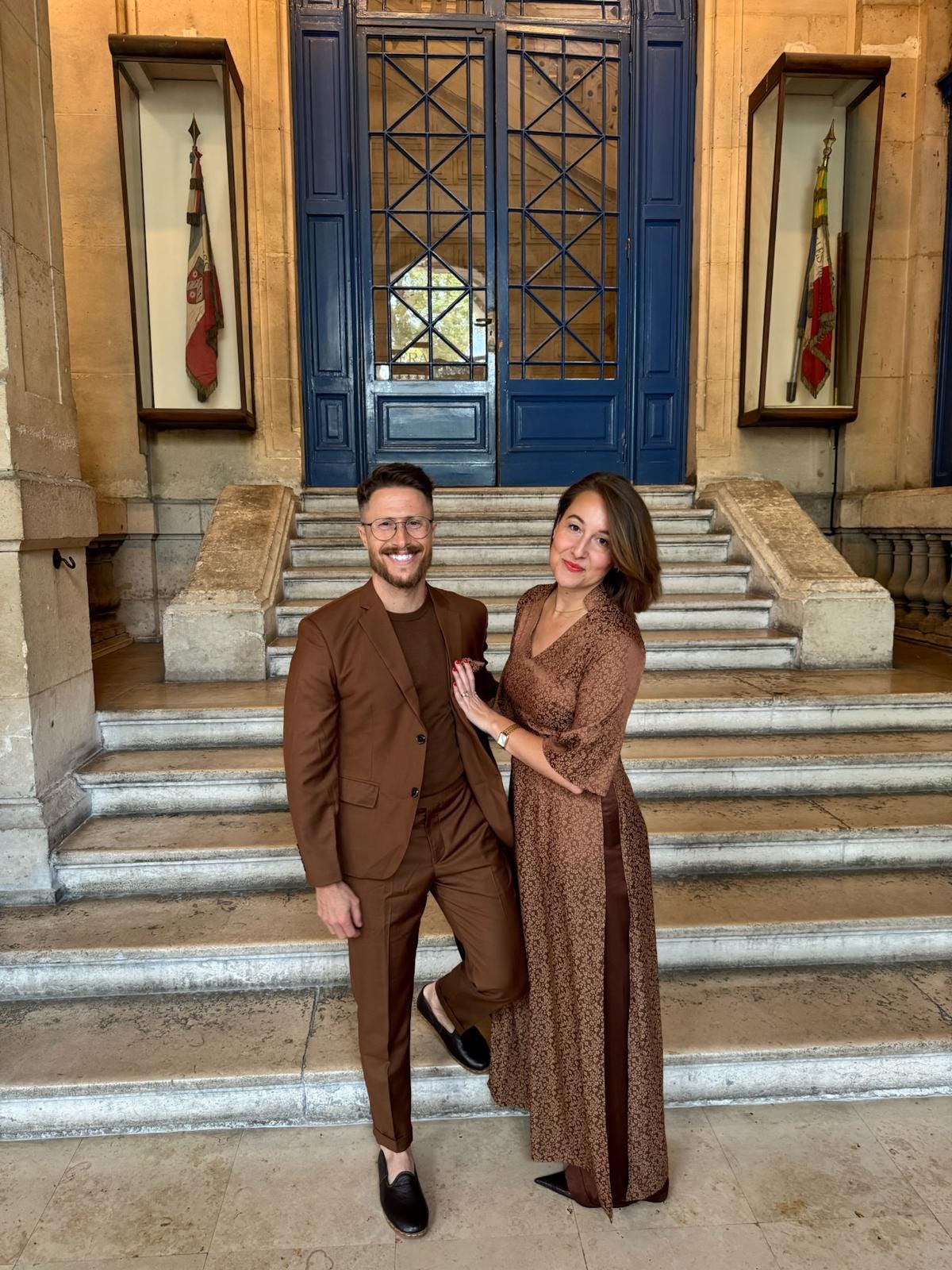 Stephen Belenky and Rachel Howze standing together on the marble steps inside the Mairie du 11ᵉ arrondissement in Paris before a wedding ceremony, both dressed in coordinated brown outfits and smiling.