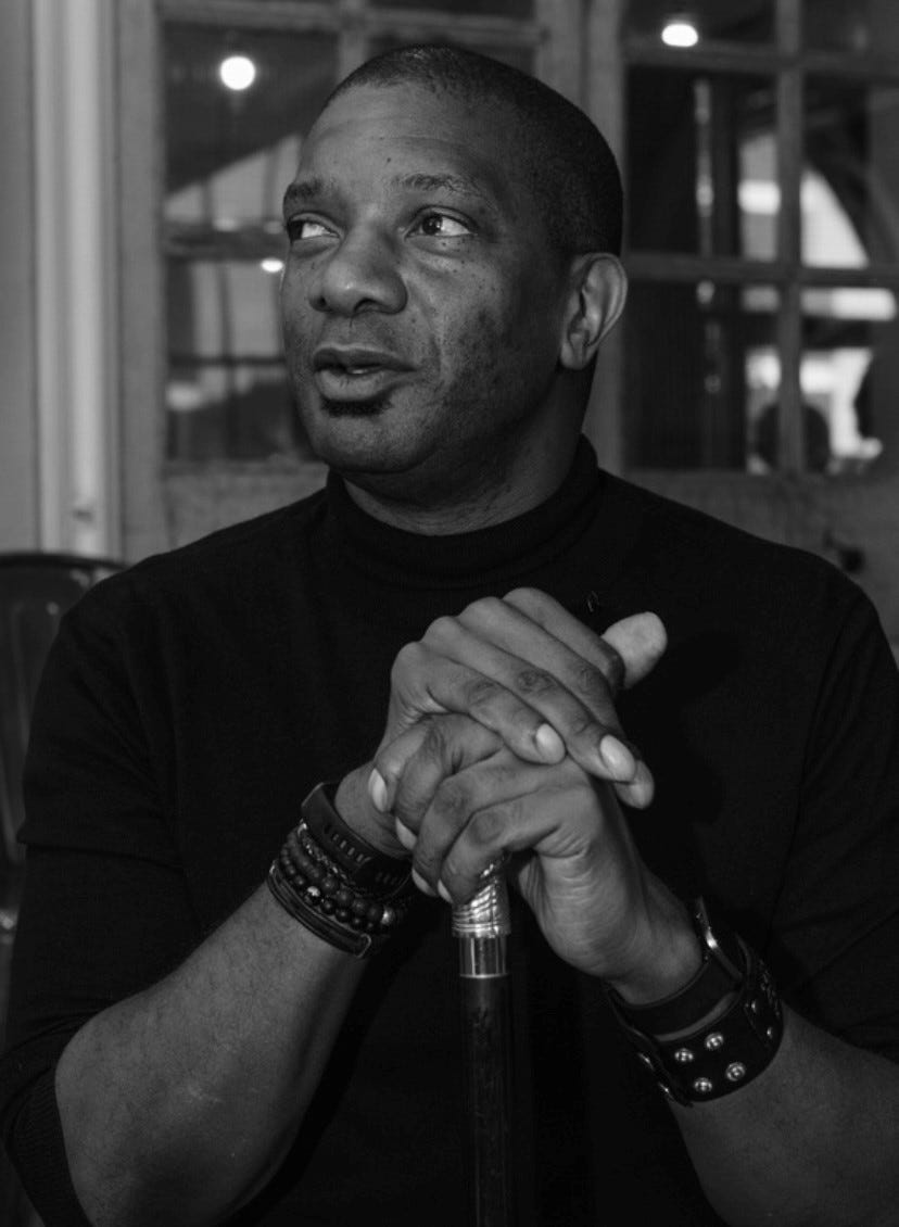 Rick Dove, a shaven headed , clean shaven black man, with his hands clasped together resting on top of his walking aid "Gregory".  Rick is wearing a watch and dressed in black polo neck and looking off camera to the left listening to someone out of frame speaking. The photo is in black and white. 