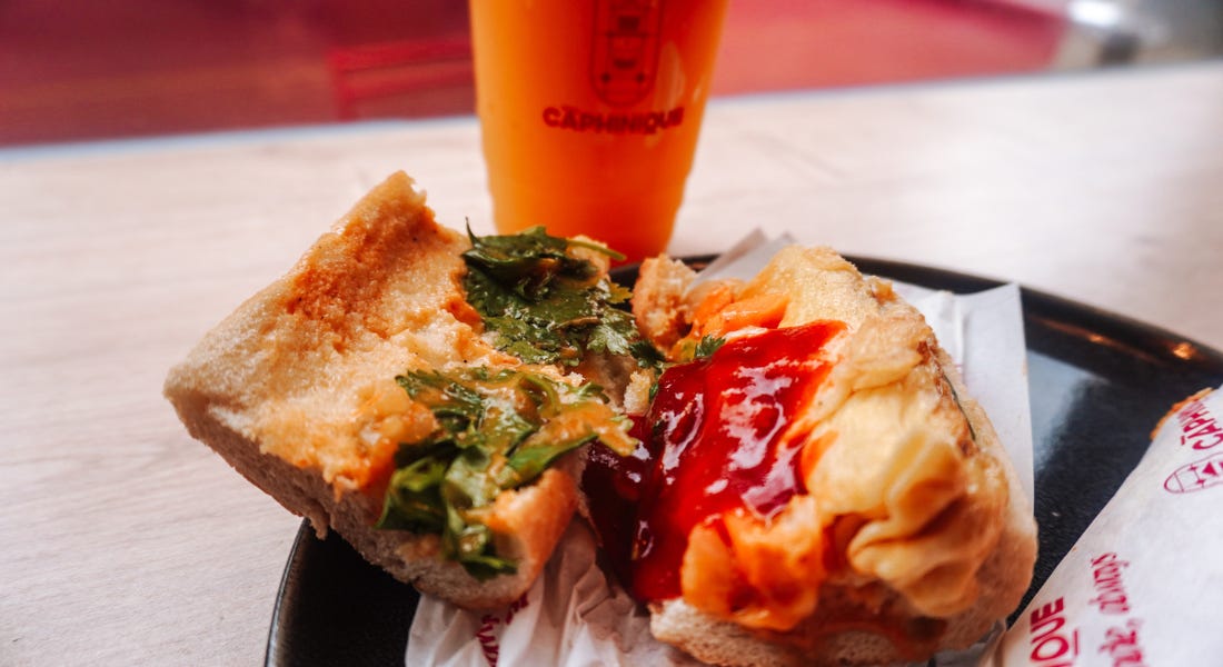 An open baguette with pickled vegetables, omelette, and sauce, with an orange drink behind it