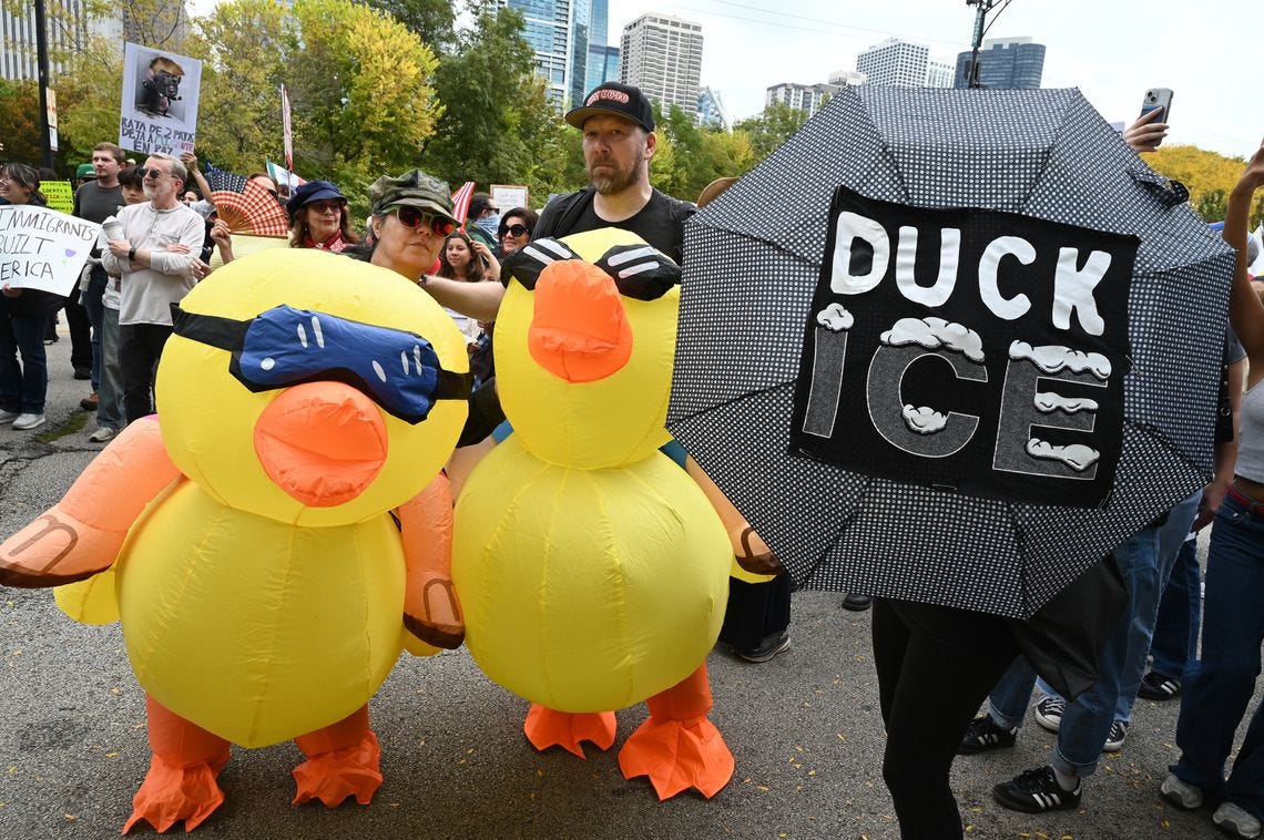 A photo shows two protesters in yellow inflatable duck costumes standing next to a person holding an open umbrella that has the words "Duck ICE" on it at a protest outdoors. 