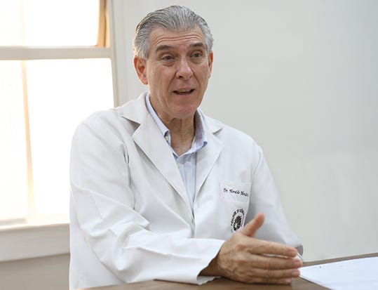 Pessoa de cabelos grisalhos usando jaleco branco com identificação, camisa azul clara por baixo, sentada à mesa em ambiente de escritório com parede branca e janela ao fundo, com a mão direita levantada em gesto de fala e expressão neutra voltada para frente. Pessoa de cabelos grisalhos usando jaleco branco com identificação, camisa azul clara por baixo, sentada à mesa em ambiente de escritório com parede branca e janela ao fundo, com a mão direita levantada em gesto de fala e expressão neutra voltada para frente.