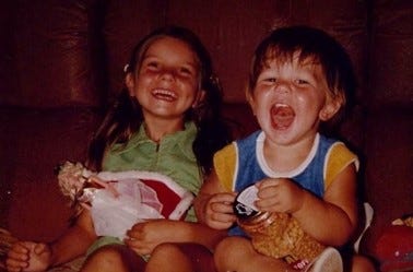 Two children sitting on a brown leather sofa in the late 70s. The girl, aged 6, haphazardly holds a Barbie doll, while the boy, age three, holds a container of peanuts. Both children have wide, open grins.