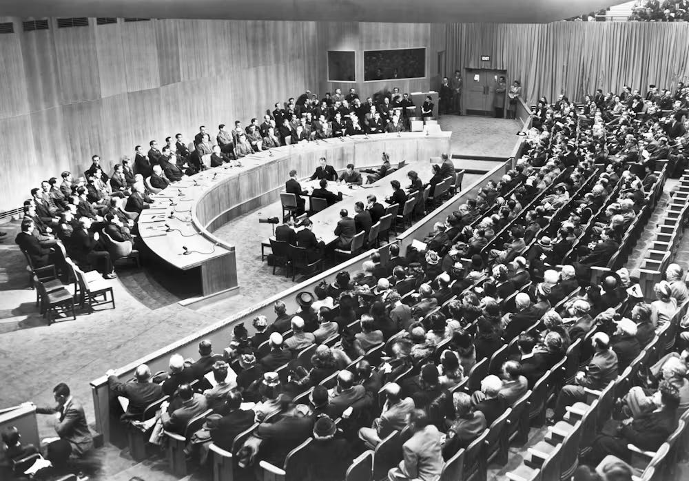 Delegates seated around a horseshoe-shaped table in a crowded hall, with rows of observers and press standing behind them, at an early meeting of the United Nations Security Council. Delegates seated around a horseshoe-shaped table in a crowded hall, with rows of observers and press standing behind them, at an early meeting of the United Nations Security Council.