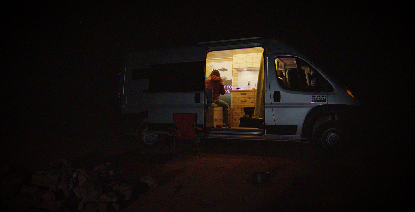 A camper van in the dark, the door open to reveal a woman on a computer with an X-Files poster on the wall.