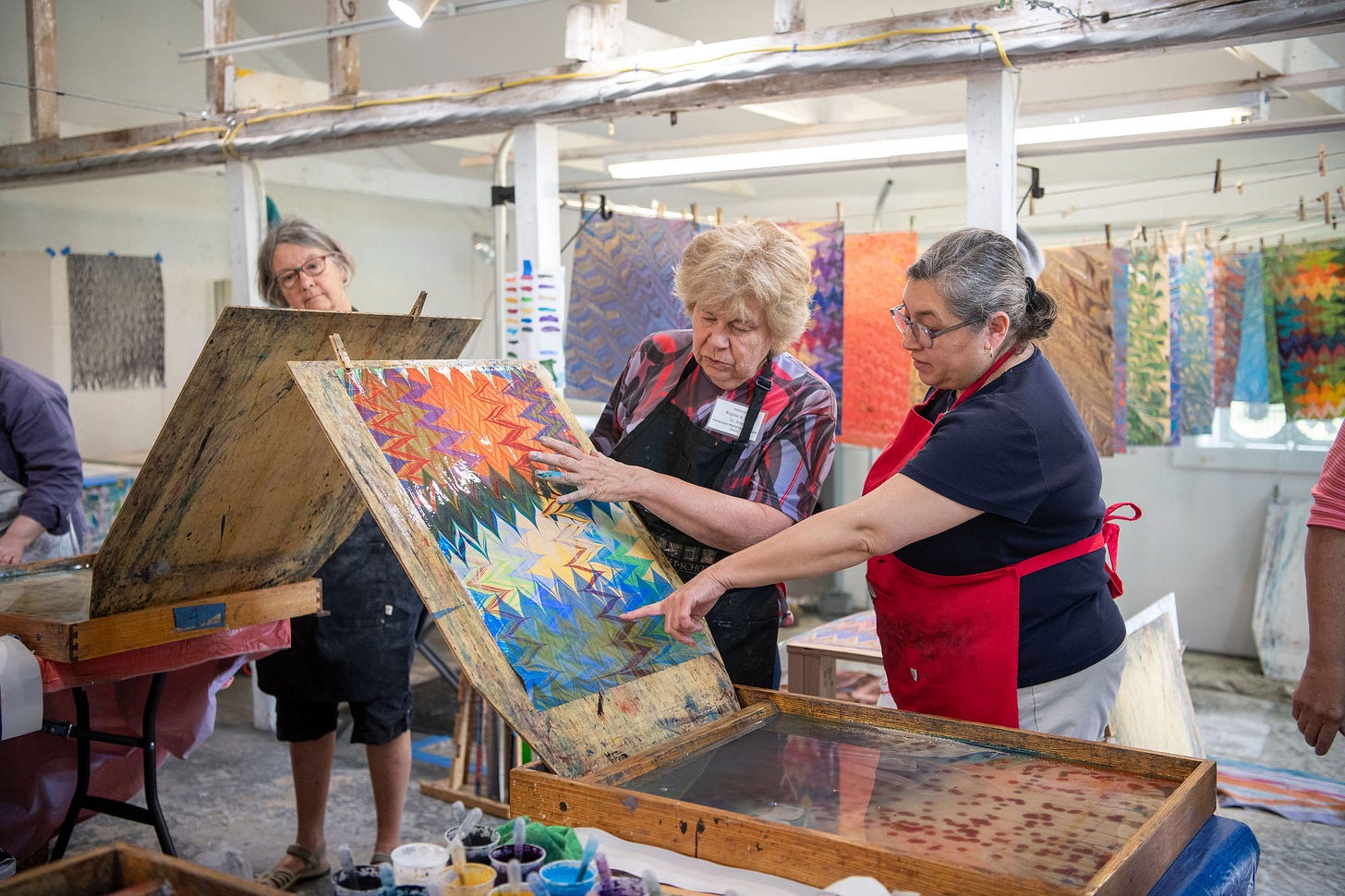 A group of women in aprons examine a colorful marbled paper during a hands-on workshop at Snow Farm.