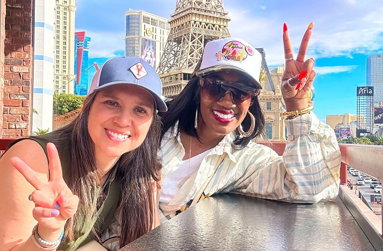 Shaunda Necole and a friend smiling and flashing peace signs while seated outdoors on the Las Vegas Strip, with the Eiffel Tower replica and Paris Las Vegas hotel in the background. A bright, joyful moment highlighting friendship and community in Las Vegas.
