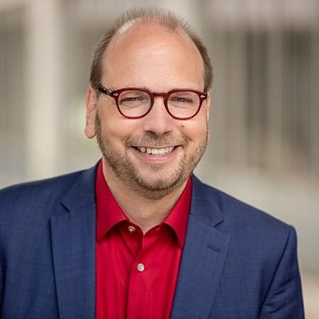 Headshot of Klaus Eck, a white man in his 50s, smiling, with glasses, a red shirt and a dark blue suit
