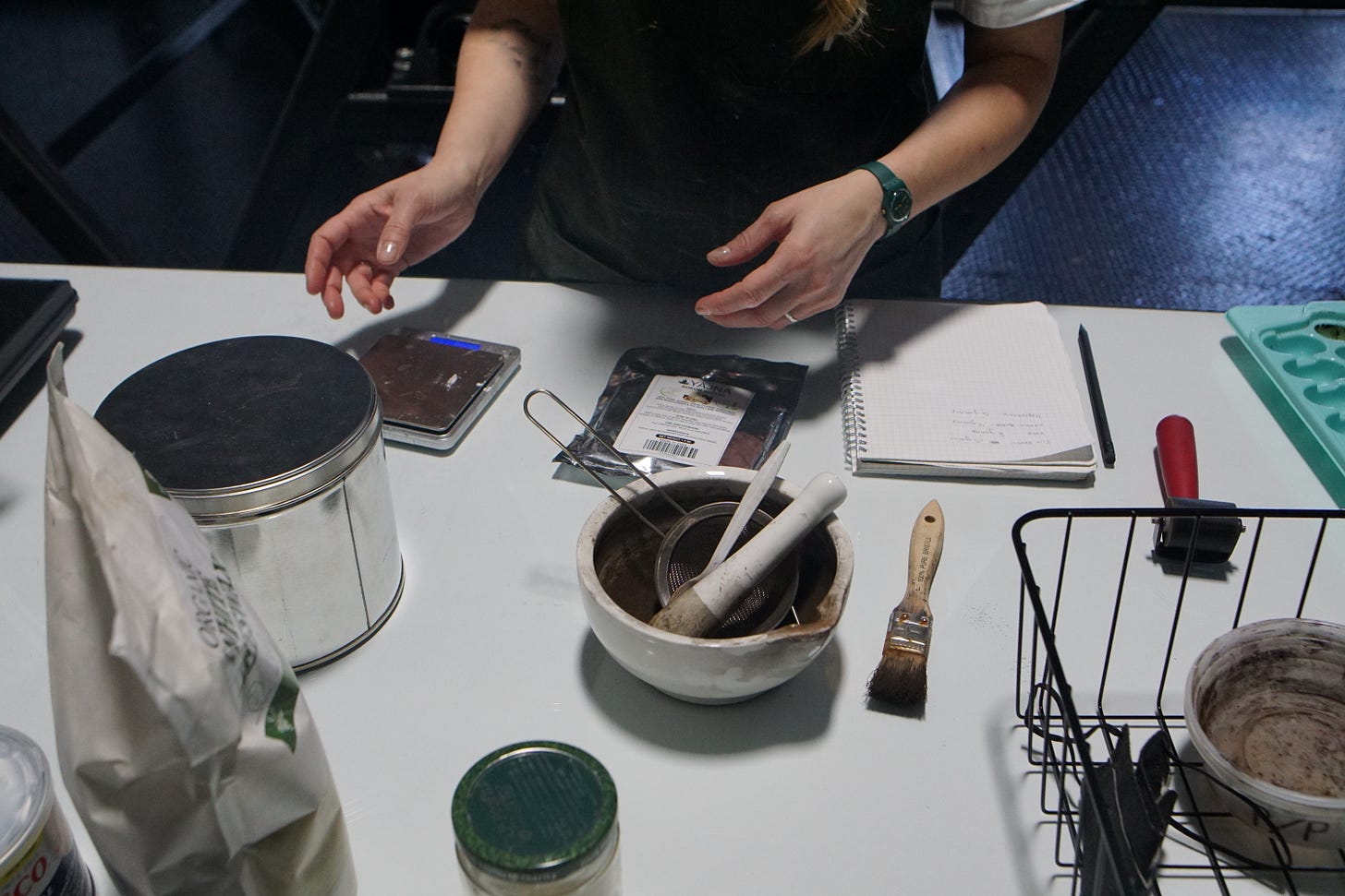 Hands seem about to reach for something on white glass work surface. On surface, rests a variety of bowls and containers.