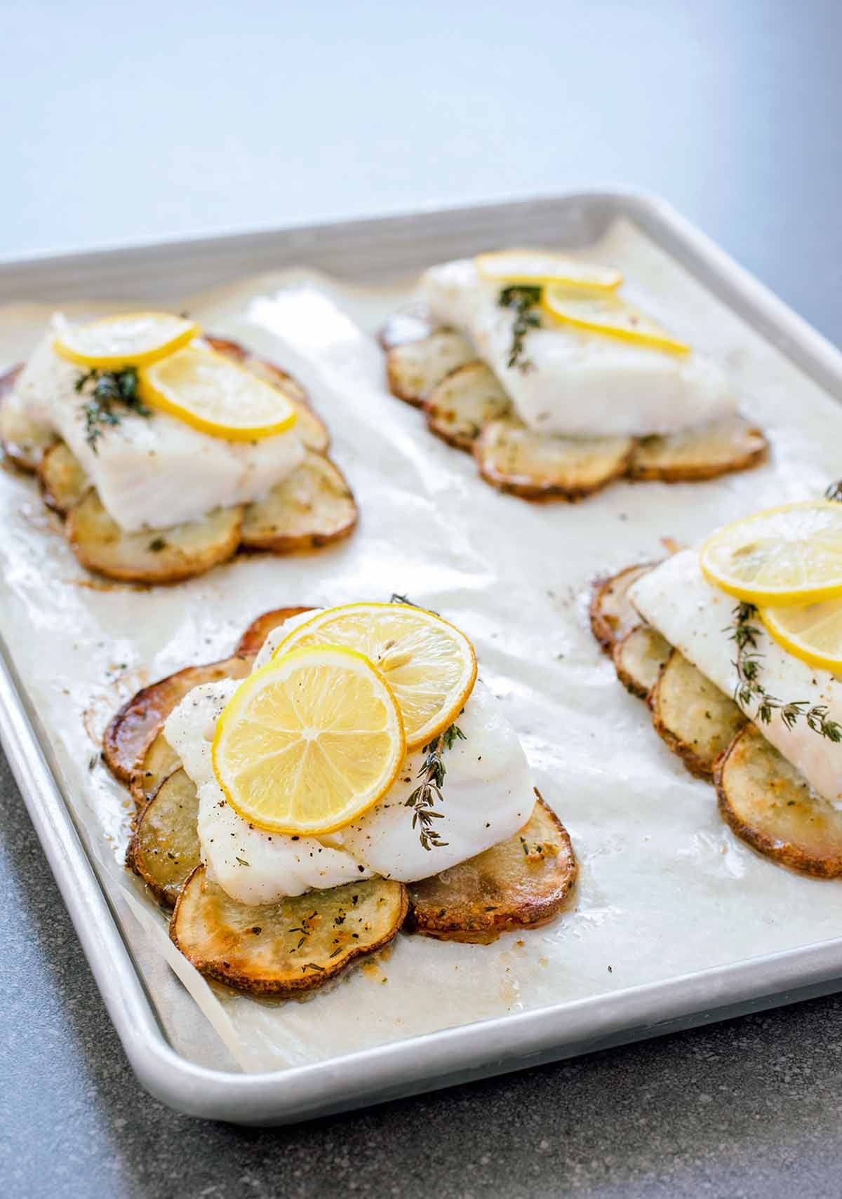 Flaky roasted cod fillets served on a bed of tender sliced potatoes with fresh herbs and bright lemon wedges on a baking sheet.