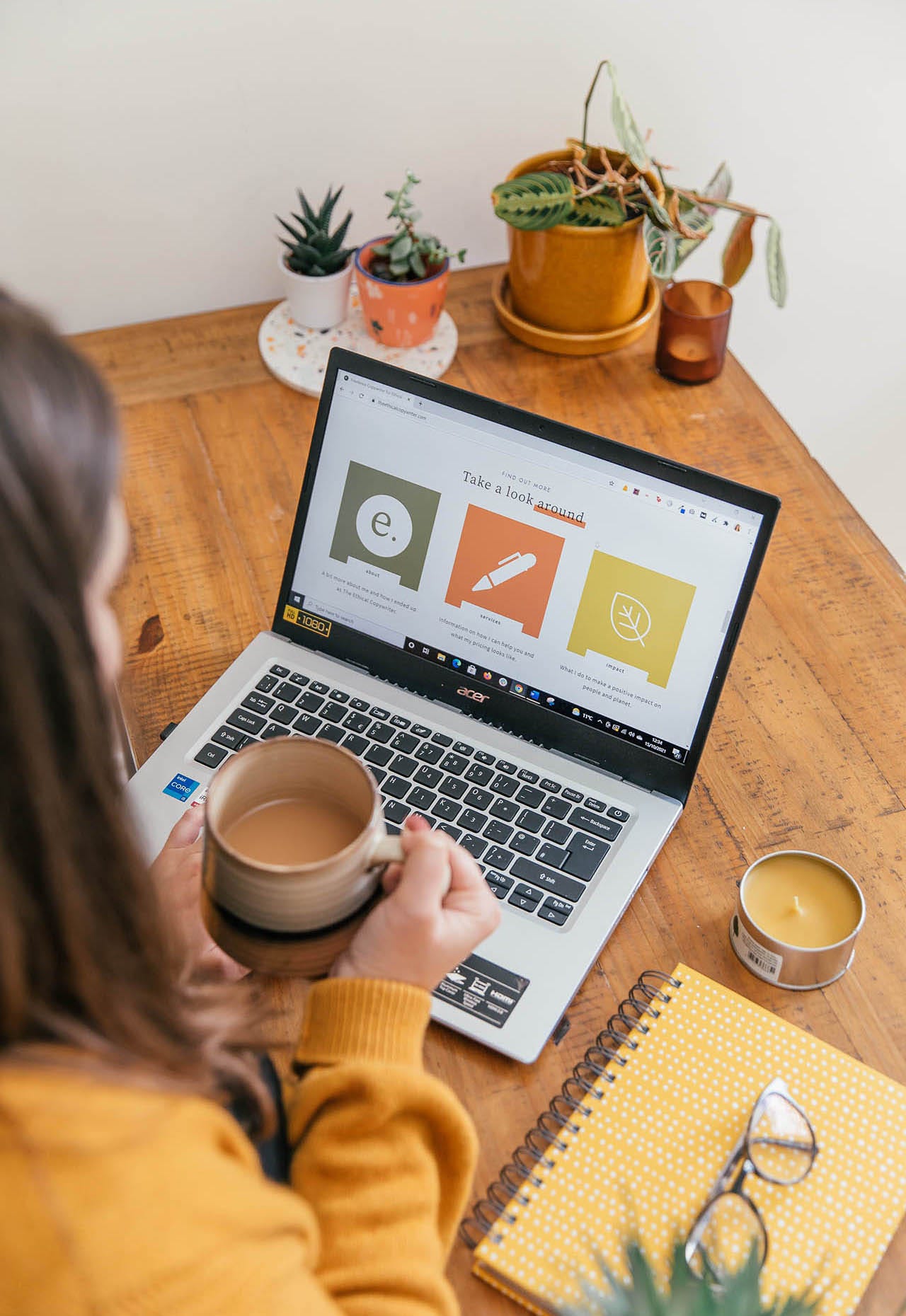 Rachel sits at her desk, holding a cup of coffee. She is looking at her laptop, which has a page from her website, theethicalcopywriter.com on the screen. The photo is taken from over her shoulder, and the full laptop screen is visible. Beside her on the desk is a yellow notebook and candle, and her glasses sit on top of the notebook. 