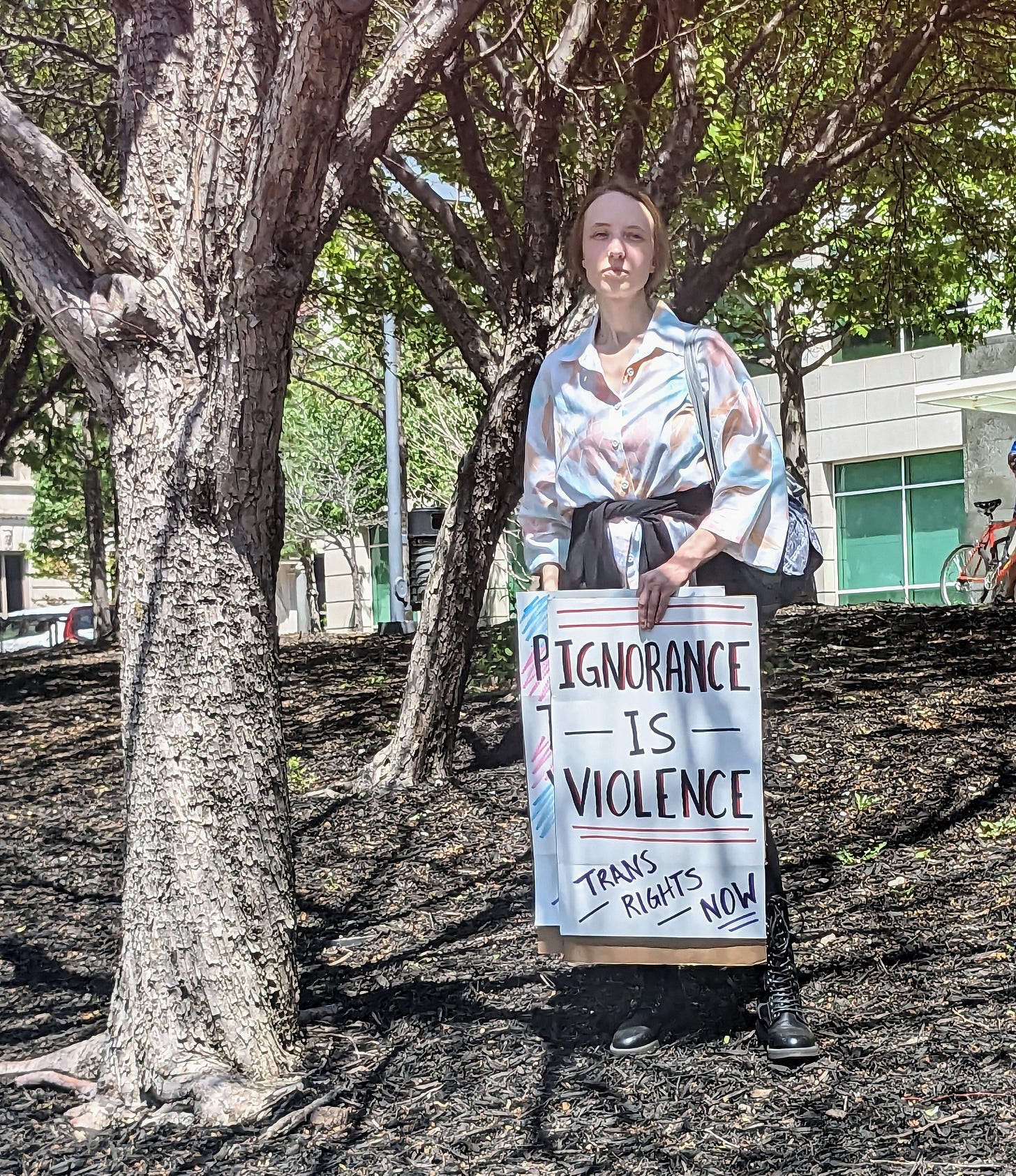 A photograph of someone standing beneath a tree, holding a sign that reads "Ignorance is Violence. Trans Rights NOW!"