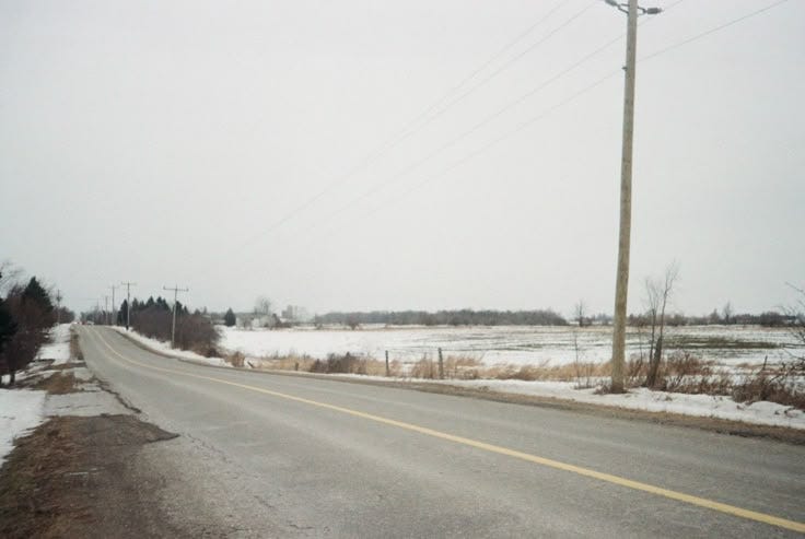 This may contain: an empty road with snow on the ground and power lines in the distance near some trees