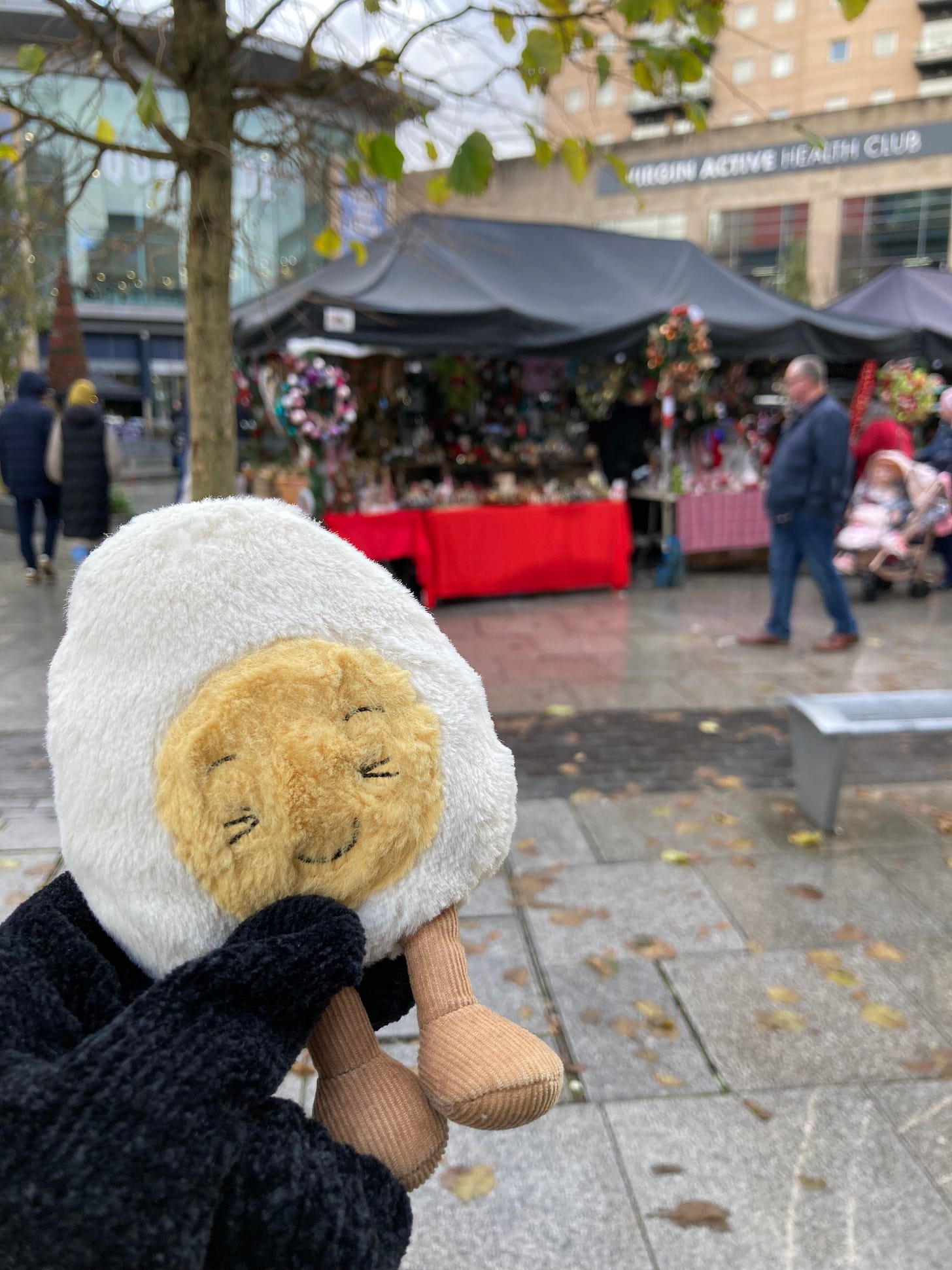 Photo of Dippy the soft toy egg in front of an outdoor christmas market, with stalls selling wreaths. It's Manchester so of course it is raining.