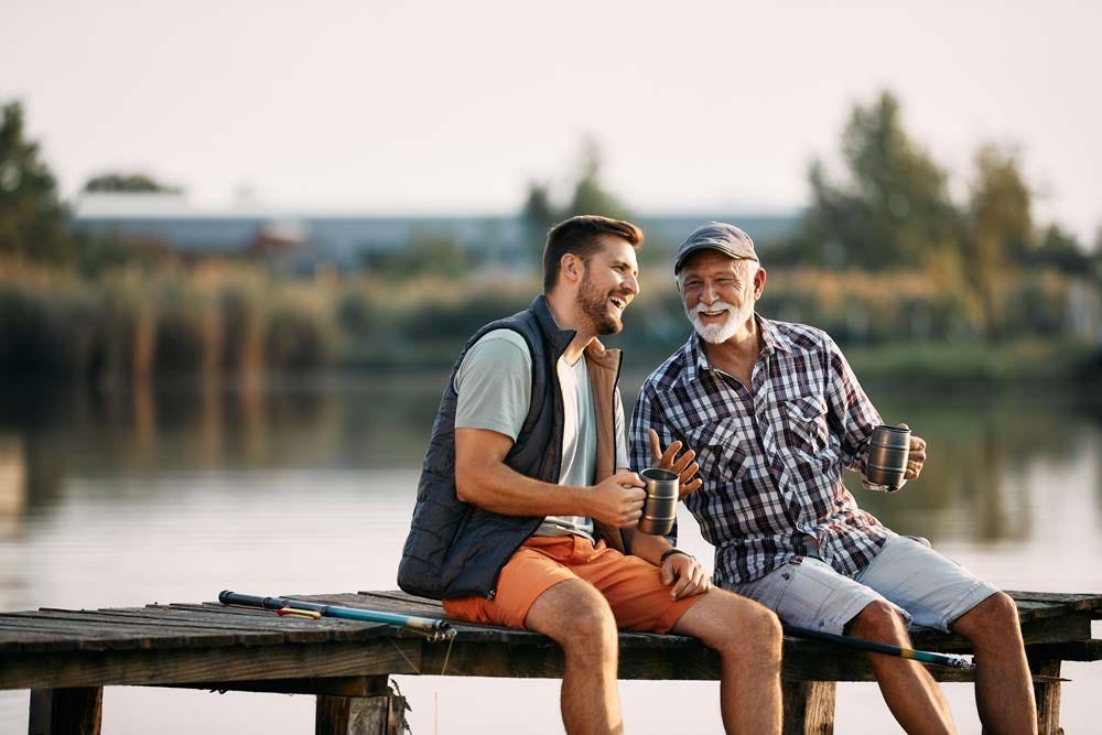 Image of an elder and his adult son enjoying a discussion while sitting out on a lakeside pier.