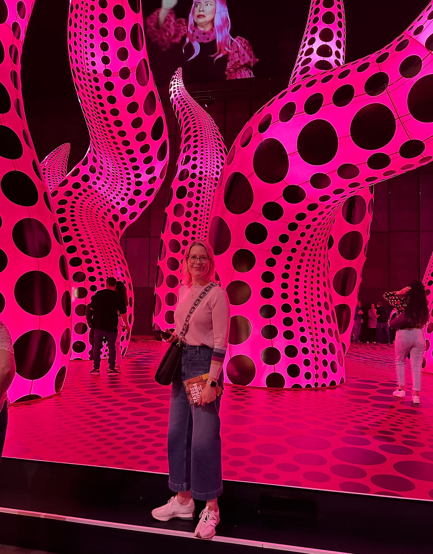 A woman stands in front of large inflatable pink tentacles with black spots on them on a raised pink platform. A video screen with a woman with pink hair is visible in the background (Yayoi Kusama).