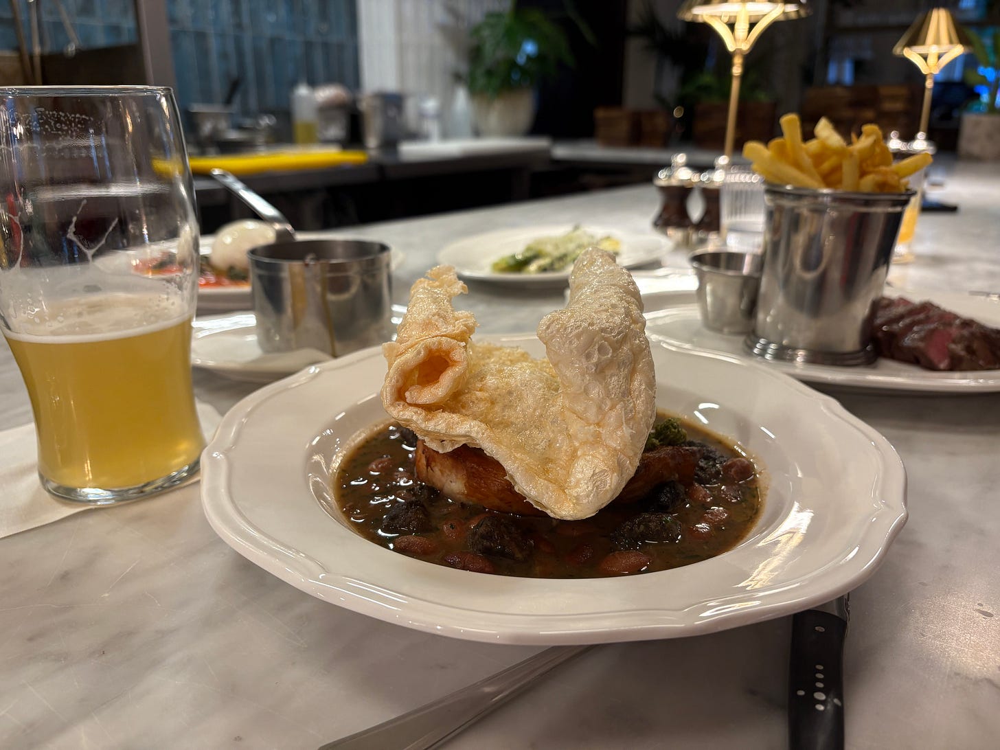 A dark-coloured stew sits in the foreground with a generous curl of 'crackling' on top, and a half-drunk pint of beer A dark-coloured stew sits in the foreground with a generous curl of 'crackling' on top, and a half-drunk pint of beer