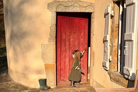 Three doors at the château: an old dark wooden door hung with a paper fairy garland, a red painted door in a stone surround with a small girl reaching for the handle, and a large arched wooden double door where two children stand dressed as a fairy and a wizard.