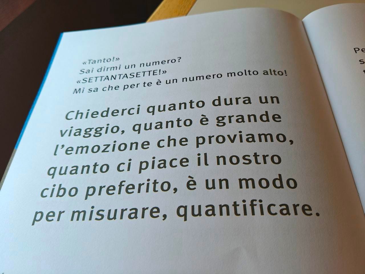 Una pagina della parola QUANTO delle Scatoline di effequ, con questa scritta: "Chiederci quanto dura un viaggio, quanto è grande l'emozione che proviamo, quanto ci piace il nostro cibo preferito, è un modo per misurare, quantificare". La pagina è appoggiata su un tavolo di legno bianco sporco e nell'angolino in alto a sinistra si intravede un pezzetto di muro in ombra.
