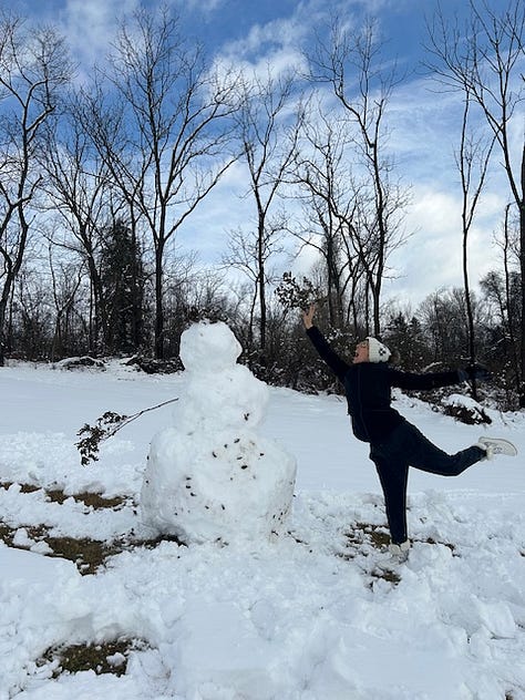 Woman makes a snow angel. Woman in a beanie cap, black jacket, and pink aviator glasses smiles in front of snow. A giant snow person hi-fives a lesbian with it's twig arm.