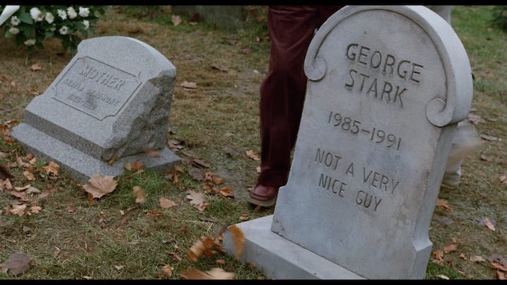 Close-up of a cemetery gravestone that reads: “GEORGE STARK, 1985–1991, NOT A VERY NICE GUY,” next to another stone labeled “MOTHER.” Fallen autumn leaves scatter across the grass.
