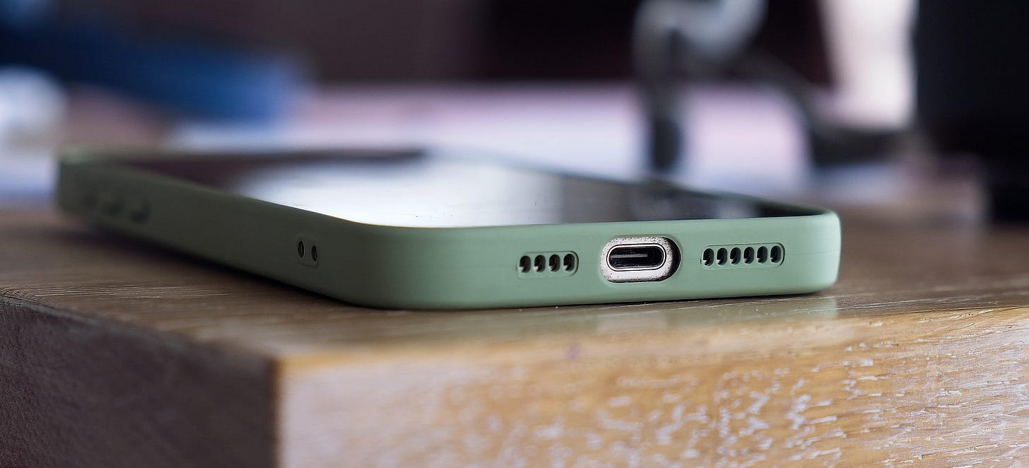 Close-up photograph of a modern smartphone in a muted green case resting on the edge of a wooden table, with the charging port and speaker grilles in sharp focus against a softly blurred background Close-up photograph of a modern smartphone in a muted green case resting on the edge of a wooden table, with the charging port and speaker grilles in sharp focus against a softly blurred background