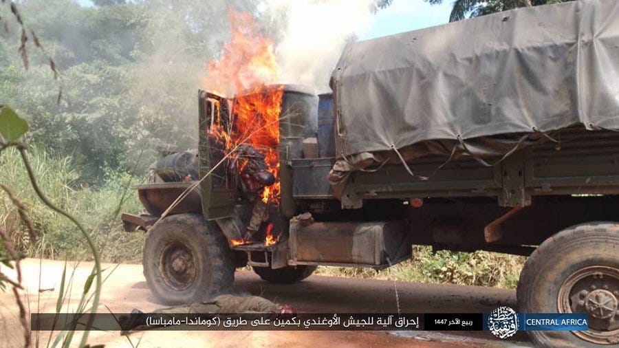 A military truck with a canvas cover is engulfed in flames on the drivers side door and cab area, with thick black smoke billowing upwards, positioned on a dirt road surrounded by green vegetation and trees in a forested setting, Arabic text overlay present.