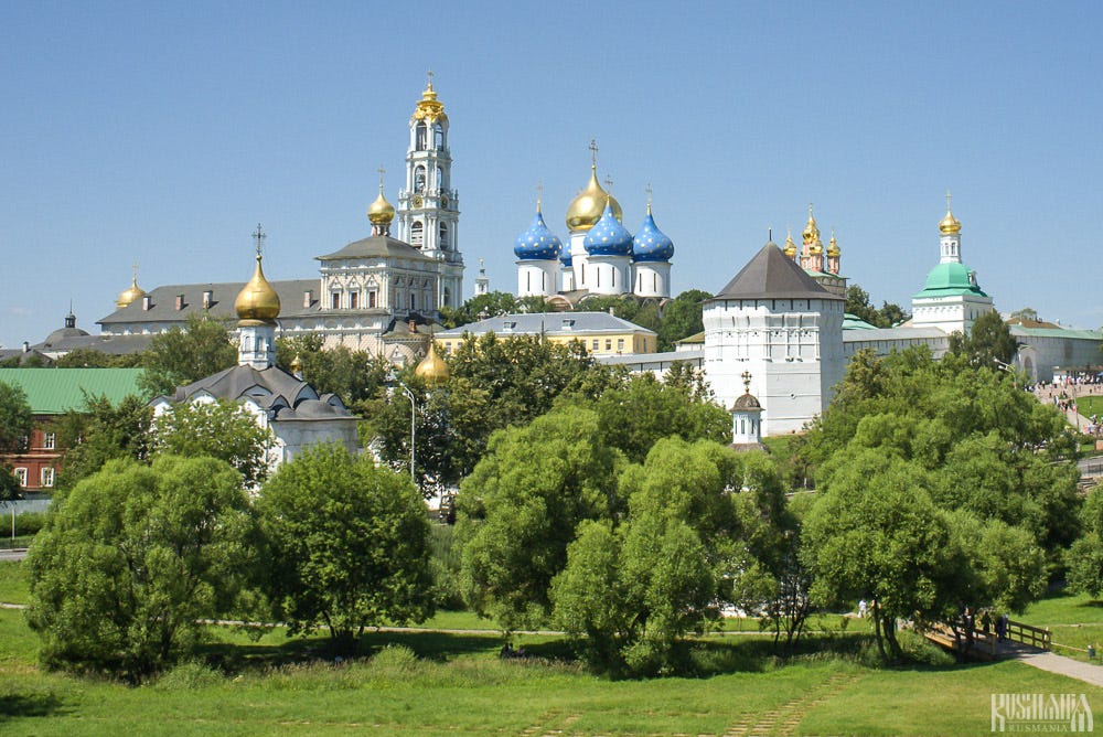 View across a park of white monastery buildings topped with gold and blue star-patterned onion domes and a tall white bell tower, framed by trees.