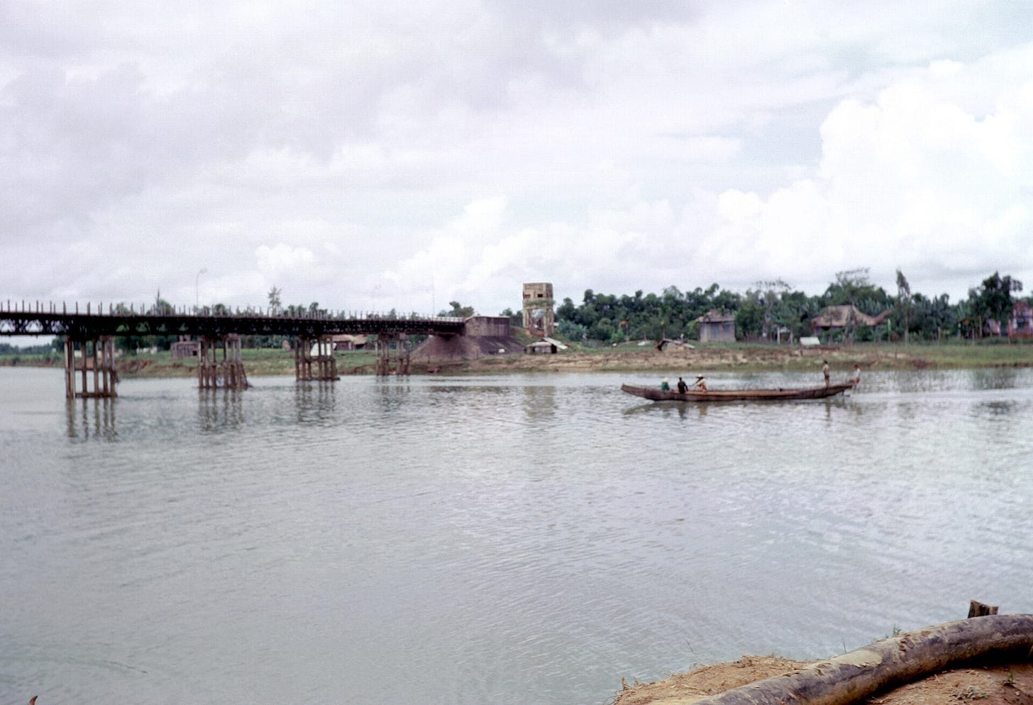 A long low bridge on concrete piers crossing a wide calm river, with a small boat in the foreground and trees on the far bank. A long low bridge on concrete piers crossing a wide calm river, with a small boat in the foreground and trees on the far bank.