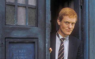 A young, slim redheaded man (Mark Strickson, playing Turlough in "Doctor Who") in the doorway of a blue police box (TARDIS).