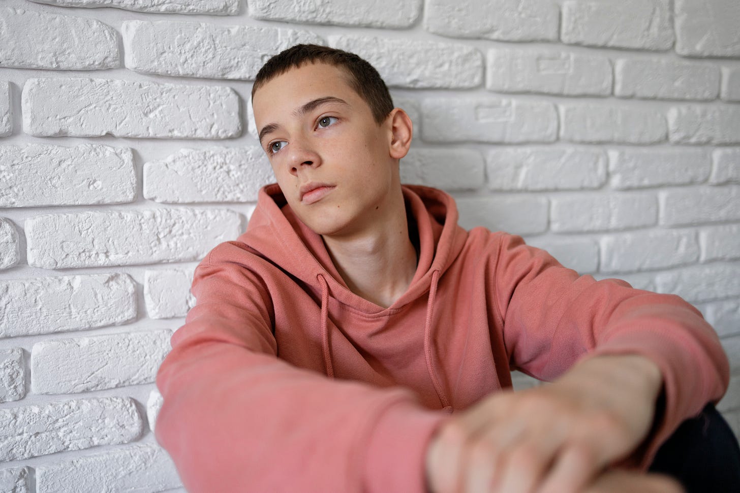 Sad-looking boy wearing a hoodie sits on the floor, leaning against a white brick wall.